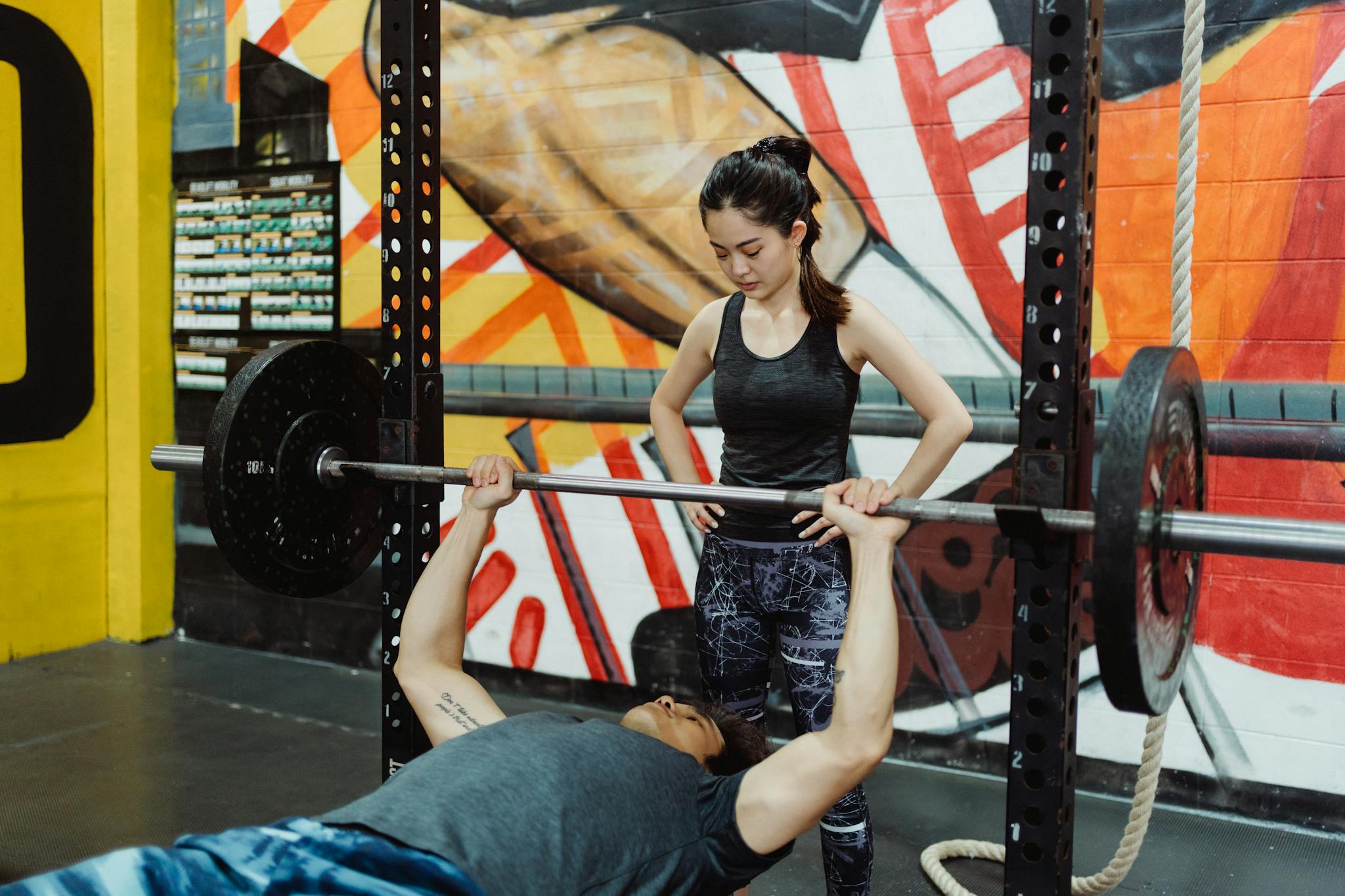 A woman coaching a man during a bench press exercise in a vibrant gym setting.