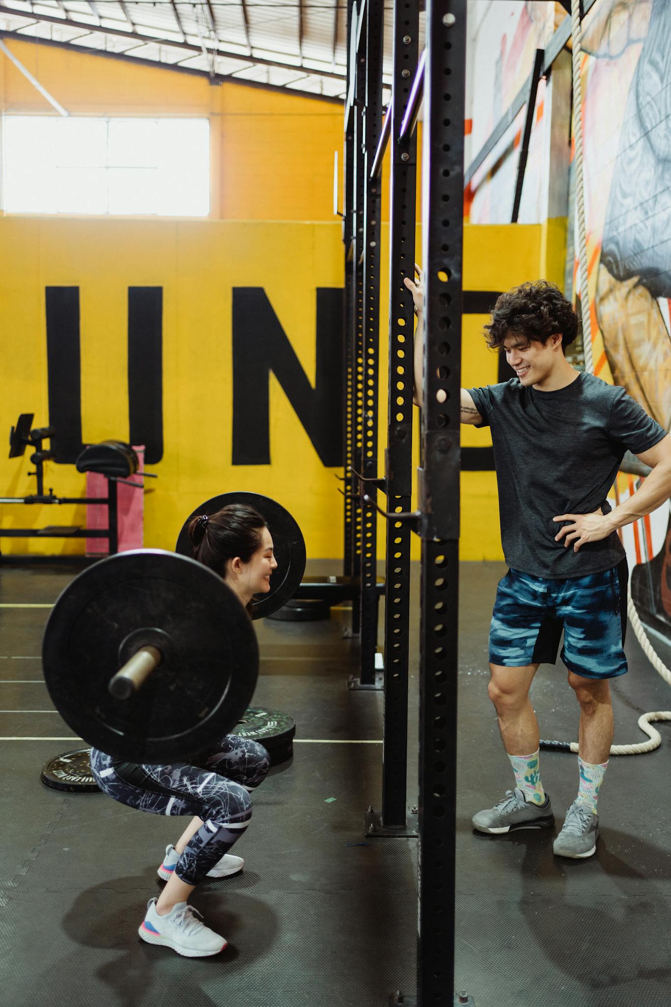 A woman lifts weights with a partner supervising in a vibrant indoor gym.