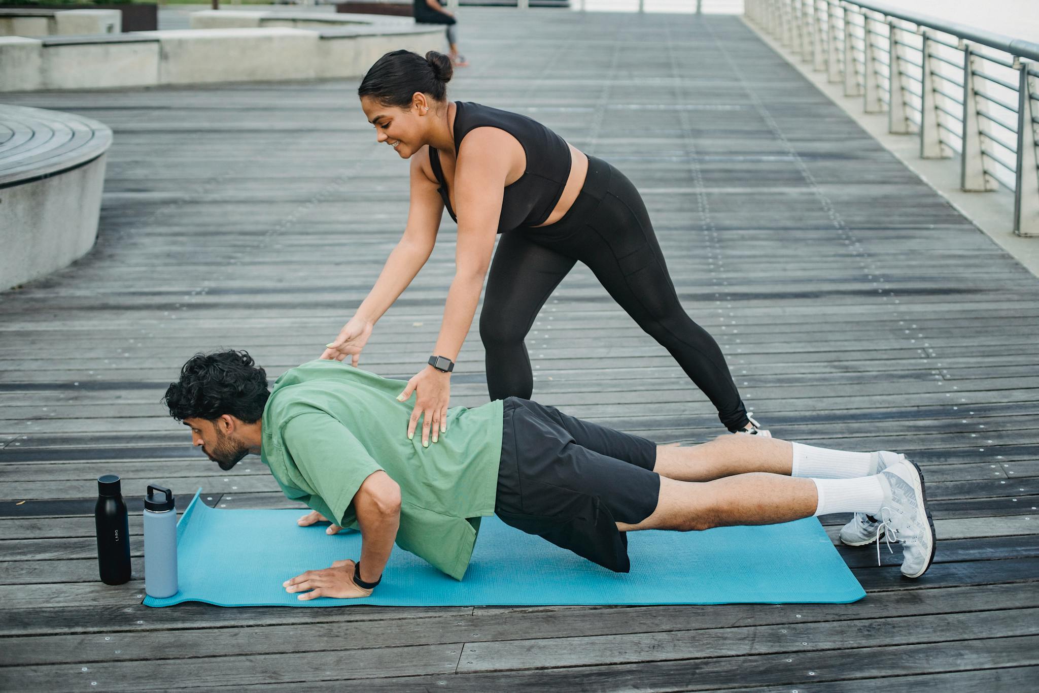 Man doing push-ups with assistance from a trainer on a boardwalk during summer.